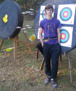 Young male archer standing in front of his target. He has a robin hood, eg. one arrow has skewered the shaft of the first arrow which is in the gold ring.