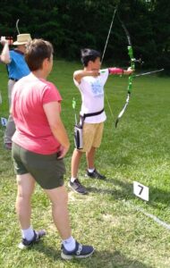 Coach Allison is standing behind a young Olympic recurve archer and watching his form. His recurve bow (with sight and stabilizer bar) has a green riser and his shooting hand is anchored carefully under his chin