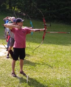 Shot of a number of archers on the shooting line. The archer in the foreground is a right handed shooter at full draw with an Olympic Recurve bow (red riser with sight and stabilizer bar.) His shooting hand is anchored under his chin.