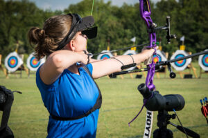 A WRA archer competing at a national tournament. She is a right hander shooting an Olympic recurve with sights and stabilizers. Her scope is in the foreground for reviewing her arrows after each shot.