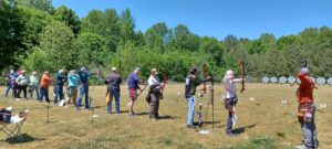 An archery field with targets 60 yards down range. On the shooting line are both men and women in the act of shooting. Some shoot olympic recurve, others barebow and compound bows. 