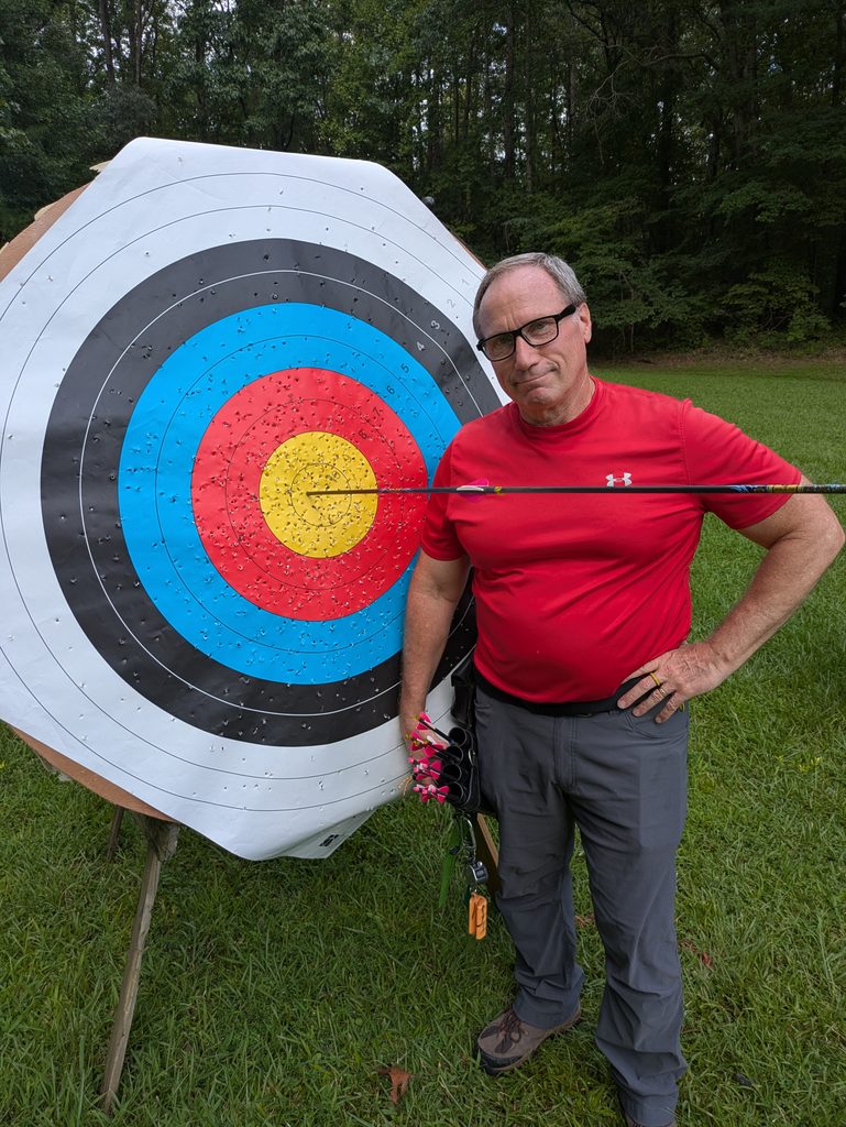 A frowning man in a red shirt standing next to an archery target with one arrow in the center and another arrow sticking out the back of the first arrow.