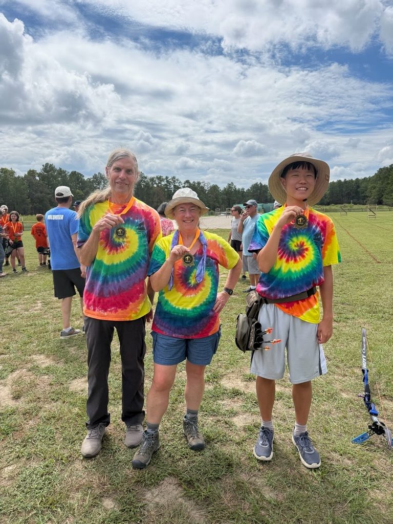 3 archers in festive tie-dyed t-shirts smiling and holding their gold medals. In the background other people are milling around an open field with trees behind and overhead a sky of mixed clouds and streaks of blue.